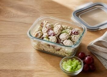 Chicken salad with red grapes in a glass storage container beside its lid, with a small bowl of diced celery and red grapes on a wooden counter