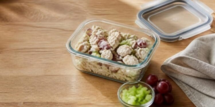 Chicken salad with red grapes in a glass storage container beside its lid, with a small bowl of diced celery and red grapes on a wooden counter