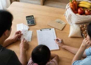Parents and a young child at a wooden kitchen table with grocery receipts, a calculator, a shopping list on a clipboard and an envelope of cash, beside a cloth bag of carrots, apples and bananas, a bag of rice and a carton of eggs