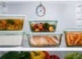 Glass containers of leftover soup, rice, roasted vegetables, sliced chicken and pasta on a refrigerator shelf, with a dial thermometer hanging above