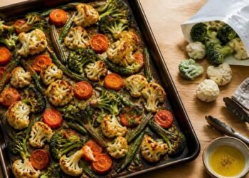 Sheet pan of roasted broccoli, cauliflower, carrot slices and green beans with browned edges, next to an open bag of frozen florets, metal tongs, a small bowl of olive oil and a linen towel on a wooden counter