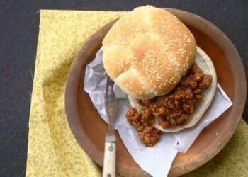 Two sloppy joe sandwiches on toasted buns on a wooden board, next to a slow cooker filled with saucy ground beef and a wooden spoon, with a small bowl of pickle slices