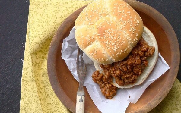 Two sloppy joe sandwiches on toasted buns on a wooden board, next to a slow cooker filled with saucy ground beef and a wooden spoon, with a small bowl of pickle slices